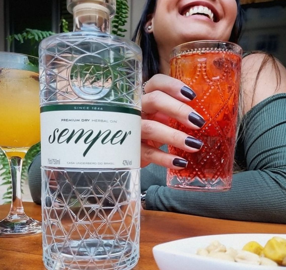 Post da Semper Gin: mulher sorrindo com um drink vermelho ao lado da garrafa